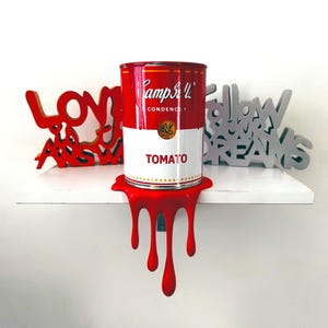 Puede incluir: Una obra de arte de una lata de sopa Campbell's Tomato roja y blanca. La lata se asienta sobre una base roja que gotea como pintura. La palabra "TOMATO" es visible en la lata. Palabras decorativas de madera "LOVE" y "DREAMS".