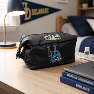 May include: A black toiletry bag featuring the embroidered text CMS in blue and yellow, along with a blue and yellow logo of a bird mascot. The bag has a textured fabric finish and a zipper closure with a pull tab on the side, presented as a functional accessory.