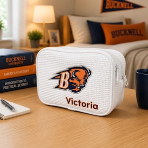 May include: A white rectangular zippered pouch with a waffle-textured fabric. The front features an embroidered orange and navy blue bison mascot logo with the letter B, and the name Victoria embroidered in orange below the mascot logo.