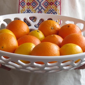 Puede incluir: Un cuenco de fruta de cerámica blanca con un diseño de celosía contiene una variedad de naranjas y limones. El cuenco está siendo sostenido por una persona que lleva una camisa blanca con un patrón bordado de colores.