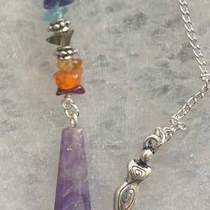 May include: A purple amethyst pendulum with a silver chain and a silver charm with a spiral design. The pendulum is adorned with a series of smaller stones in various colors, including amethyst, blue, green, orange, and red.
