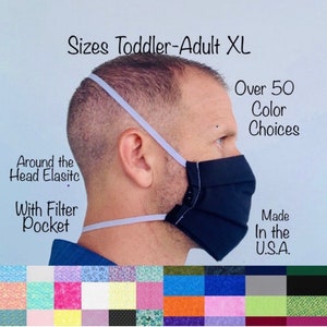 May include: A side view of a person wearing a dark blue face mask with a white elastic band around the head. The mask has a filter pocket and a button closure. The text "Sizes Toddler-Adult XL", "Over 50 Color Choices", "Around the Head Elastic", "With Filter Pocket", and "Made In the U.S.A." is visible in the image. A row of fabric swatches in various colors and patterns is shown at the bottom of the image.