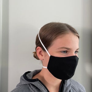May include: Black face mask with white elastic ear loops and a white elastic strap around the back of the head. The mask is made of a soft, dark fabric. The person is wearing a gray hooded sweatshirt.