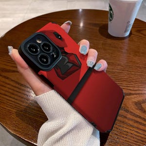 Puede incluir: Funda de teléfono roja con un diseño gráfico en negro y rojo. La funda tiene un borde negro y una superficie texturizada. El diseño parece ser un personaje de cómic. La funda del teléfono está sobre una mesa de madera.