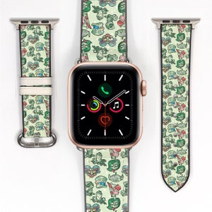 May include: A green, white and pink Apple Watch band with a repeating pattern of cartoon characters. The band is shown on a gold Apple Watch.