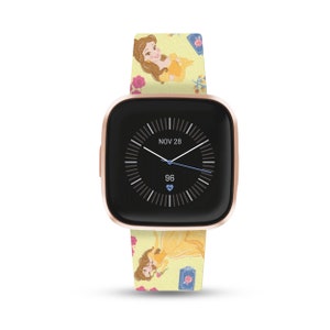 K&ouml;nnte beinhalten: Eine ros&eacute;goldene Fitbit Smartwatch mit einem gelben und rosafarbenen Armband mit Belle aus Disneys Die Sch&ouml;ne und das Biest. Das Zifferblatt der Uhr zeigt das Datum "NOV 28" und eine Herzfrequenz von 96.