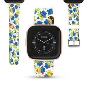 Puede incluir: Un smartwatch de color oro rosa con una banda blanca que presenta un patrón repetitivo de extraterrestres verdes de la franquicia Toy Story. La esfera del reloj muestra la fecha "NOV 28" y la hora 96.