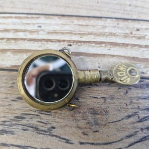 Puede incluir: Espejo compacto vintage de latón con forma redonda y un pequeño mango. El espejo tiene una superficie reflectante y está unido a un mango con la palabra "INDIA" grabada. El compacto está sobre una superficie de madera.