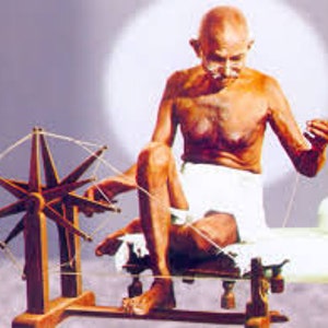 May include: A man sits on a white cloth, spinning a wooden spinning wheel with a white thread. He is shirtless and has a white cloth wrapped around his waist. The spinning wheel is on a wooden stand with a white thread attached to it.