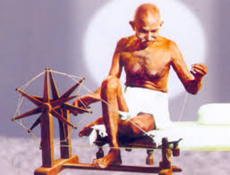 May include: A man sits on a white cloth, spinning a wooden spinning wheel with a white thread. He is shirtless and has a white cloth wrapped around his waist. The spinning wheel is on a wooden stand with a white thread attached to it.