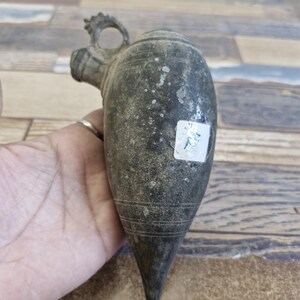 May include: A vintage, dark metal plumb bob with a rounded top, a small handle, and a pointed bottom. The plumb bob has a textured surface and a small sticker on the side. The object is held in a hand.