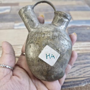 May include: A small, antique silver-colored metal pitcher with a handle. The pitcher has a textured surface and two spouts. A small white label with the handwritten text "H4" is attached to the pitcher.