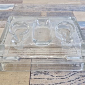 May include: A clear glass ashtray with three recessed areas. Two circular areas with a curved notch are on either side of a rectangular area. The ashtray is rectangular with a thick, clear border. The ashtray sits on a wooden surface.