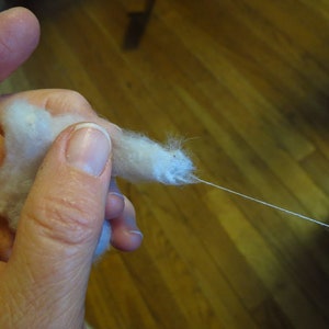 May include: A hand holding a clump of white cotton fiber with a thin white thread extending from it.