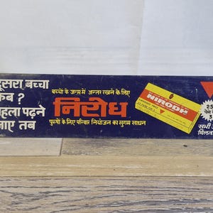 May include: Vintage rectangular sign with Hindi text and a yellow box labeled "NIRODH." The sign is dark blue with white and red text, and a wooden surface below. The sign promotes family planning.
