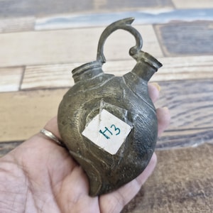 May include: Antique brass-colored metal container with a leaf-like shape, featuring a curved handle and two spouts. A small white tag with the handwritten number "H3" is attached to the front. The container is held in a hand.