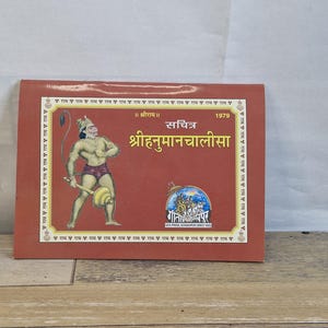 May include: A red book with gold text and illustrations. The cover features a depiction of a muscular figure holding a mace and a circular object. The title "श्रीहनुमानचालीसा" is prominently displayed, along with the year 1979.