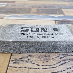 Op de afbeelding: Een rechthoekige grijze korund slijpsteen met het woord "SUN" in zwarte blokletters. De tekst "CORUNDUM SHARPENING STONE FINE & COARSE" is eronder gedrukt. De steen ligt op een houten oppervlak.