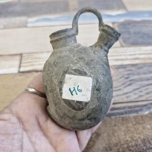 May include: A small, weathered, gray metal container with a handle and two spouts. A small white label with the number "H6" is affixed to the front. The container is held in a person's hand.