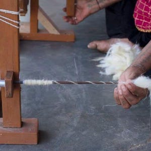 May include: A person is spinning white yarn on a wooden spinning wheel. The yarn is being pulled from a ball of white yarn and is being spun into a thin thread.
