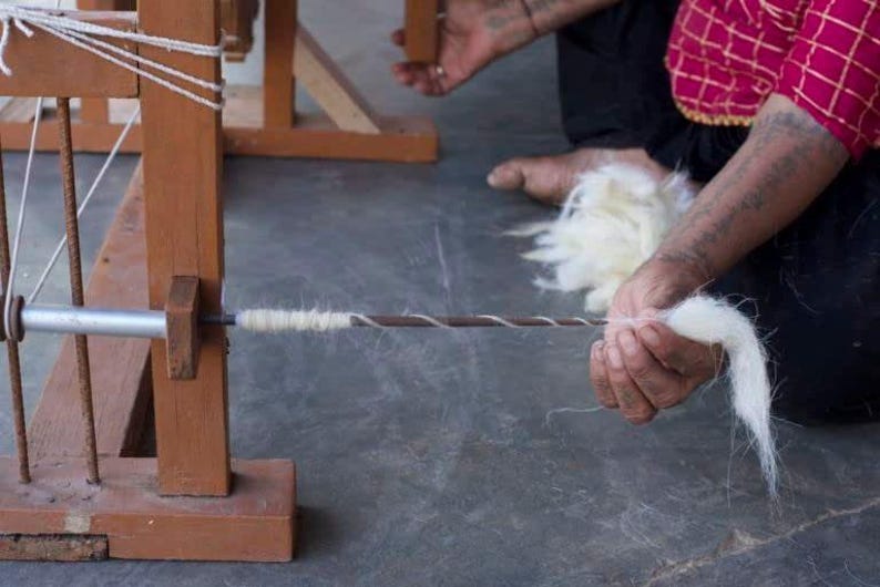 May include: A person is spinning white yarn on a wooden spinning wheel. The yarn is being pulled from a ball of white yarn and is being spun into a thin thread.