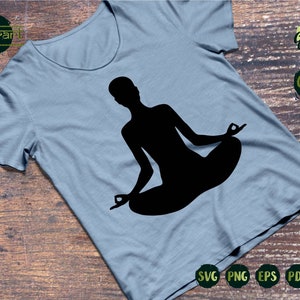 May include: A black silhouette of a person sitting in a yoga pose on a light blue t-shirt. The t-shirt is on a wooden surface. The text "Example Cutting Files" is in the upper right corner of the image.