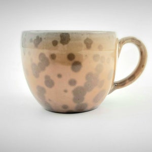 Könnte beinhalten: Eine Keramik-Kaffeetasse mit rundem Körper und Henkel. Die Tasse hat eine hellpfirsichfarbene Grundfarbe mit unregelmäßigen, dunkelgrauen Flecken. Der Rand und der Henkel sind in einem dunkleren Braunton gehalten.