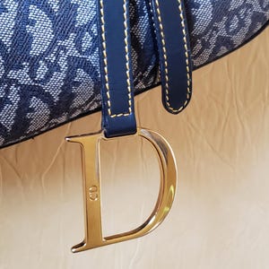May include: Close-up of a navy blue and white patterned handbag. The bag features a navy blue leather strap with gold stitching and a large gold-colored metal "D" charm with the initials "CD".