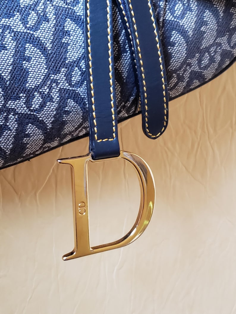 May include: Close-up of a navy blue and white patterned handbag. The bag features a navy blue leather strap with gold stitching and a large gold-colored metal "D" charm with the initials "CD".