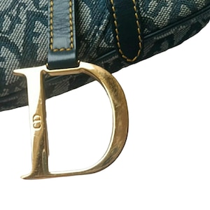 May include: Close-up of a blue and white patterned handbag with a large gold-colored metal "D" charm. The bag has a dark blue leather strap with gold stitching. The charm also has the letters "CD" on it.