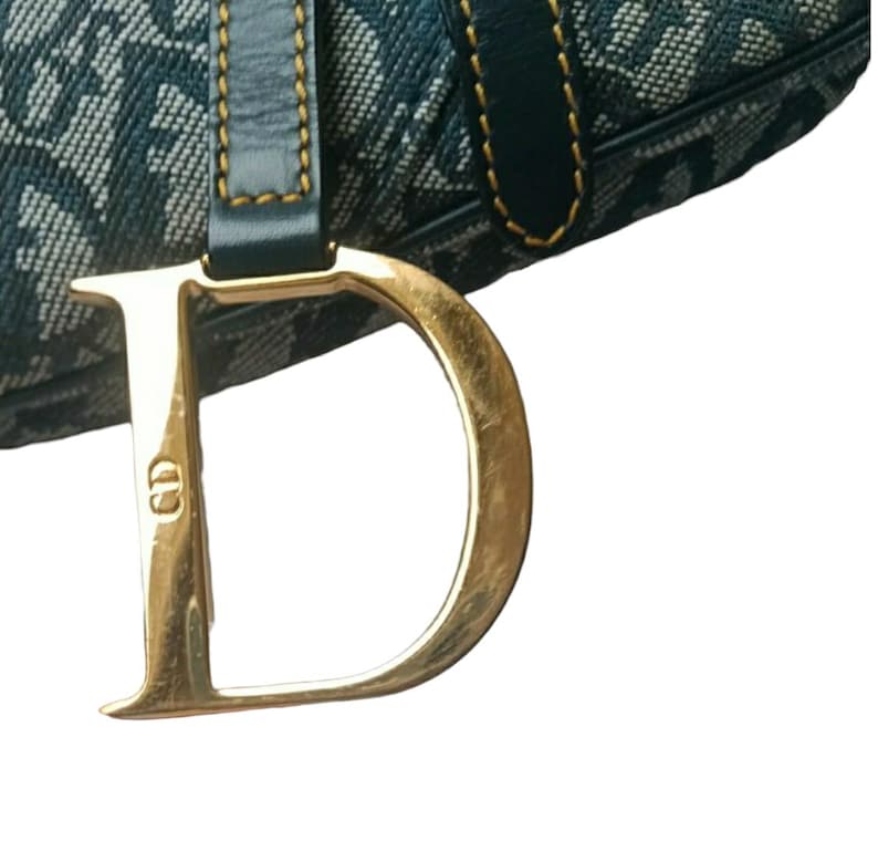 May include: Close-up of a blue and white patterned handbag with a large gold-colored metal "D" charm. The bag has a dark blue leather strap with gold stitching. The charm also has the letters "CD" on it.