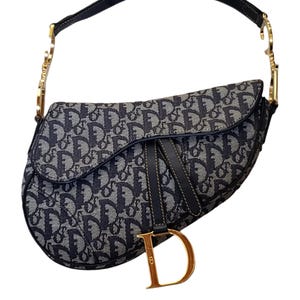 May include: A navy blue and gray saddle bag with a repeating pattern. The bag has a black leather strap and gold-colored hardware. A gold-colored "D" charm hangs from the front.