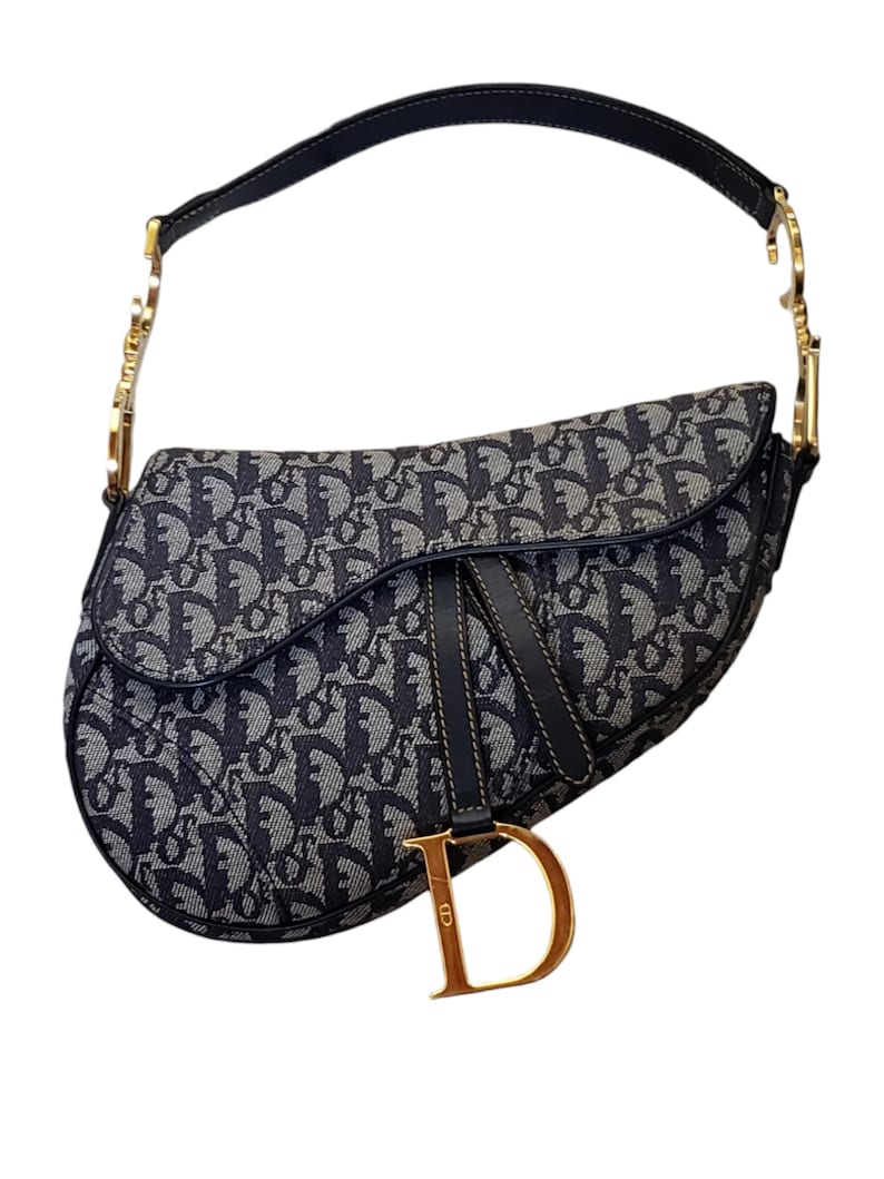 May include: A navy blue and gray saddle bag with a repeating pattern. The bag has a black leather strap and gold-colored hardware. A gold-colored "D" charm hangs from the front.
