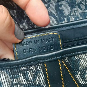 May include: Close-up of a navy blue handbag with a repeating white pattern. The bag features a leather tag with "MADE IN ITALY" and the code "01RU 0073" stitched in gold thread. The bag's seams are also highlighted with gold stitching.