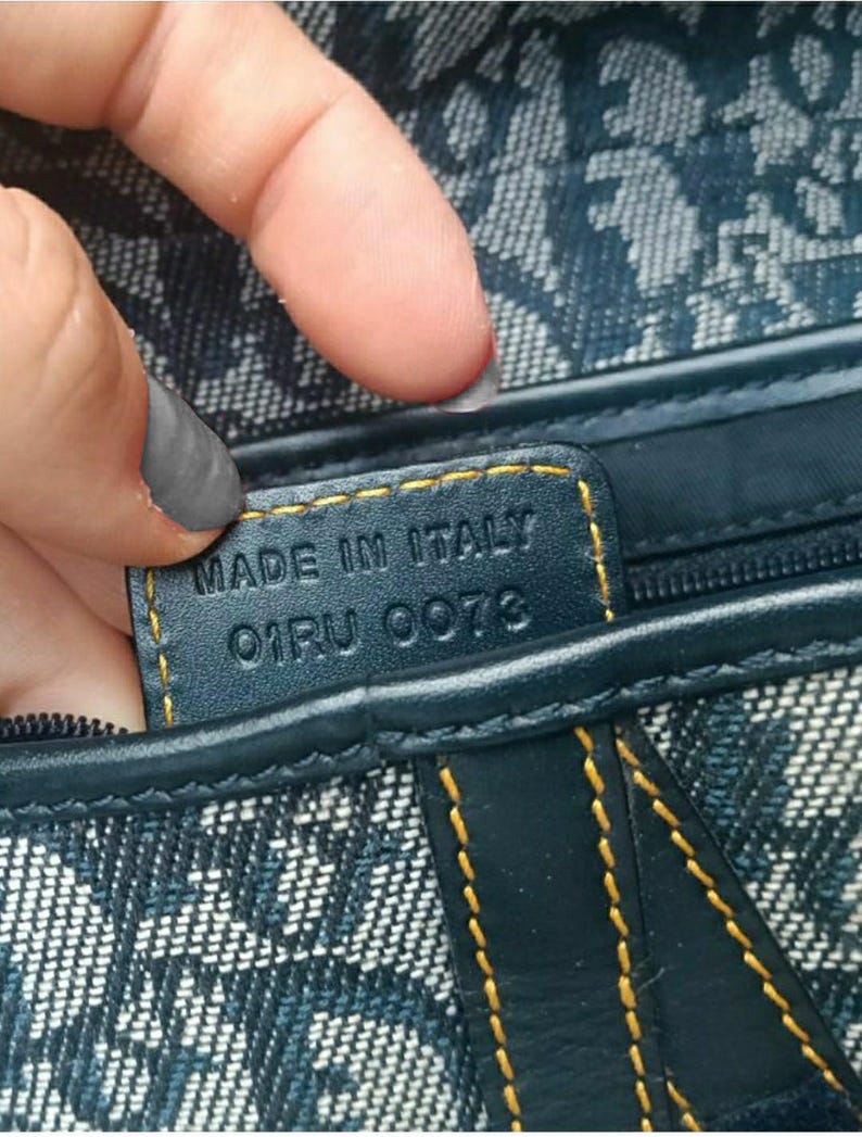 May include: Close-up of a navy blue handbag with a repeating white pattern. The bag features a leather tag with "MADE IN ITALY" and the code "01RU 0073" stitched in gold thread. The bag's seams are also highlighted with gold stitching.