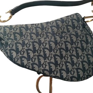 May include: A blue and gray saddle bag with a repeating pattern. The bag has a curved shape, a top handle, and a shoulder strap. The hardware is gold-colored.