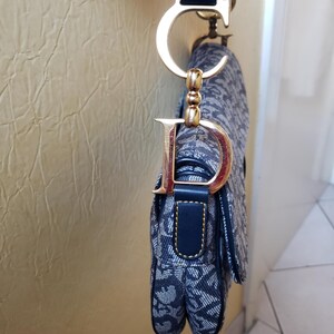 May include: A navy blue and white patterned handbag with a gold-tone metal "D" charm and a matching strap. The bag features a flap closure and a structured design, showcasing a classic and elegant style.