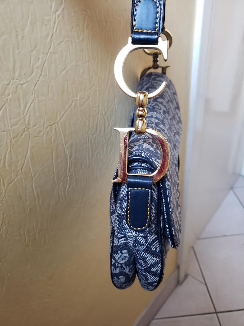 May include: A navy blue and white patterned handbag with a gold-tone metal "D" charm and a matching strap. The bag features a flap closure and a structured design, showcasing a classic and elegant style.