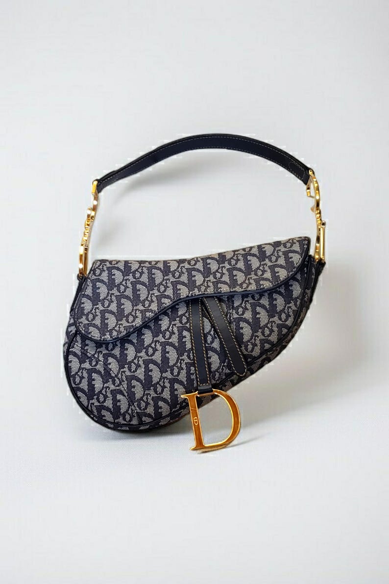 May include: A navy blue and gray saddle bag with a repeating pattern. The bag has a black handle and strap, with gold-colored hardware. A gold-colored "D" charm hangs from the front of the bag.