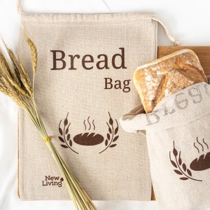 May include: Two beige linen bread bags with brown text and a bread graphic. The larger bag says "Bread Bag" and the smaller bag says "New Living". A loaf of bread is in the smaller bag.