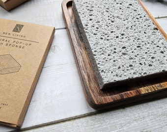 May include: A brown cardboard box with the text "NEW LIVING NATURAL POP-UP DISH SPONGE" and "Plastic Free & Compostable". The box is next to a wooden tray with a gray, porous sponge inside.