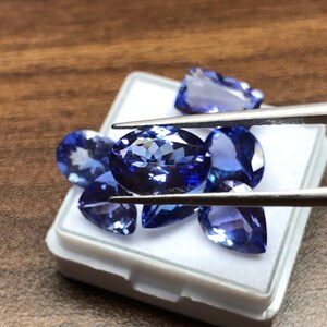May include: A collection of blue gemstones in a white box. The gemstones are oval and pear shaped. The gemstones are being held by tweezers.