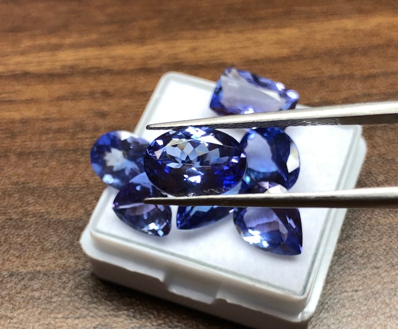 May include: A collection of blue gemstones in a white box. The gemstones are oval and pear shaped. The gemstones are being held by tweezers.