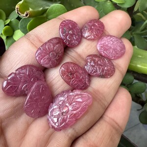 May include: A collection of carved pink gemstones, each with a floral design. The stones vary in shape, including oval and teardrop, and are held in a hand. The gemstones have a polished, translucent appearance.