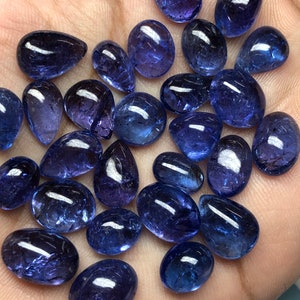 May include: A collection of polished, oval and teardrop-shaped gemstones in varying sizes. The stones are a deep, translucent blue with a glossy finish, reflecting light. The gemstones are displayed on a light-colored surface.