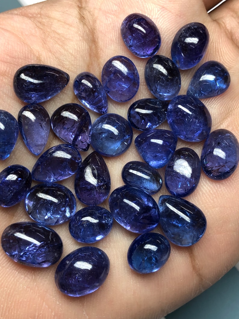 May include: A collection of polished, oval and teardrop-shaped gemstones in varying sizes. The stones are a deep, translucent blue with a glossy finish, reflecting light. The gemstones are displayed on a light-colored surface.