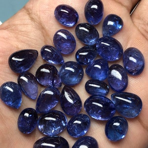 May include: A collection of polished, oval and teardrop-shaped tanzanite gemstones. The gemstones are a deep, rich blue-violet color, with a glossy, reflective surface. The stones vary in size and are displayed on a light-colored surface.