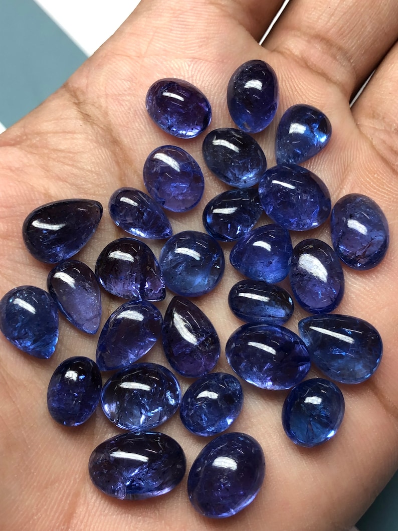 May include: A collection of polished, oval and teardrop-shaped tanzanite gemstones. The gemstones are a deep, rich blue-violet color, with a glossy, reflective surface. The stones vary in size and are displayed on a light-colored surface.