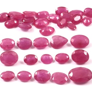 May include: A collection of faceted, oval-shaped rubies in varying sizes, arranged on a white surface. The gemstones display a deep, vibrant pink-red hue, with visible facets catching the light. The arrangement includes individual stones and small groupings.