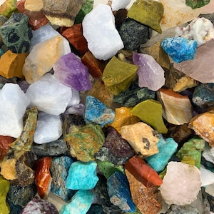 Op de afbeelding: Een verzameling van verschillende kleurrijke en getextureerde natuurlijke stenen, waaronder amethist, turquoise en jaspis. De stenen zijn in een willekeurig patroon gerangschikt, waardoor hun unieke vormen en kleuren worden getoond.